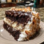 A decadent slice of Almond Joy Layer Cake featuring rich chocolate cake, creamy filling, chocolate ganache, and toasted coconut flakes.