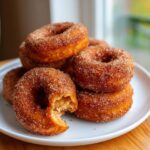 A stack of freshly made Apple Cider Donuts coated in cinnamon sugar, with one donut bitten into.