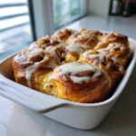 Close-up of a white baking dish filled with golden brown Apple Cinnamon Roll Bake, drizzled with white icing.