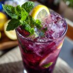 A close-up of a tall glass filled with vibrant purple Blackberry Mint Lemonade, crushed ice, fresh mint sprigs, and lemon slices.