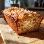 A slice of moist Coconut Macadamia Cake topped with toasted coconut flakes, resting on a wooden board.