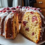 A festive Cranberry Gingerbread Bundt Cake, drizzled with white glaze and topped with cranberries. One slice is cut out, revealing the cake's texture.
