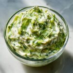 A close-up of a clear glass bowl filled with creamy Cucumber Dill Yogurt Dip, showcasing fresh dill and cucumber pieces.