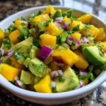 A close-up of a white bowl filled with vibrant Mango Avocado Salsa with Lime, featuring diced mango, avocado, red onion, and cilantro.
