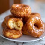 A stack of freshly made Maple Glazed Donuts, with one donut showing a bite taken out.