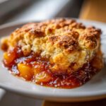 A close-up of a serving of Peach Bourbon Cobbler on a white plate, showing juicy peaches and a golden-brown biscuit topping.