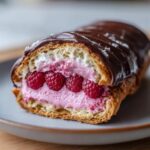 Close-up of a Raspberry Cream Éclair cut in half, revealing pink cream filling and fresh raspberries, topped with dark chocolate glaze.