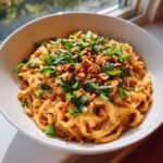 A close-up of a bowl of Spicy Thai Peanut Noodles, generously topped with chopped peanuts, green onions, and fresh cilantro.