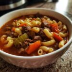 A close-up of a white bowl filled with comforting hamburger soup with macaroni, carrots, and celery.