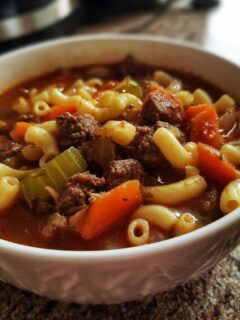 A close-up of a white bowl filled with comforting hamburger soup with macaroni, carrots, and celery.