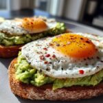 Close-up of two slices of delicious avocado toast with egg, seasoned with chili flakes and black pepper.