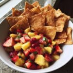 A white bowl filled with a vibrant fruit salsa made of strawberries and pineapple, served with cinnamon chips.