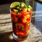 A tall glass filled with Mexican Shrimp Cocktail, featuring plump shrimp, diced avocado, cilantro, and a lime wedge.