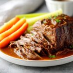 A close-up of a tender, shredded Ultimate Slow Cooker Pot Roast served with carrots and celery in rich gravy.