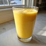 A tall glass filled with a vibrant, bright yellow Anti Inflammatory Pineapple Citrus Smoothie, sitting on a white counter near a window.