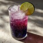 A tall glass filled with a layered Blueberry Ginger Lime New Years Mocktail, ice, and a lime wheel garnish.