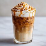 A tall glass of Caramel Oat Latte Mocktail, layered with ice, milk, caramel sauce, topped with whipped cream and cookie crumbles.