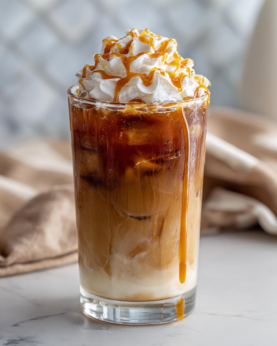 A tall glass filled with ice, layered milk, and coffee, topped with whipped cream and drizzled with caramel sauce. This is a Caramel Oat Latte Mocktail.