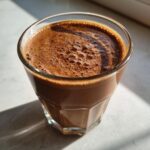 Close-up of a frothy Chocolate Espresso Smoothie in a faceted glass, with sunlight casting shadows.