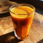 A glass of creamy Jamaican inspired vegan carrot juice with a frothy top, lit by sunlight.