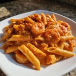 A close-up of creamy, orange-hued Cajun Pasta featuring penne noodles and seasoned shrimp on a white square plate.