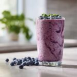 A tall glass filled with a vibrant Easy Spinach Blueberry Smoothie, topped with blueberries and spinach leaves. Fresh blueberries are scattered on the counter.
