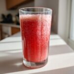 A tall glass filled with vibrant red Fresh Watermelon Cucumber Mint Juice, showing foam on top, sitting in sunlight.