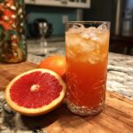 A tall glass filled with a vibrant pink Grapefruit Apple Cider Vinegar Drink and ice, next to fresh grapefruit halves.