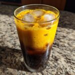 A tall glass containing a layered Honey Turmeric Iced Latte with ice cubes, showing a dark bottom layer and a vibrant yellow top layer.