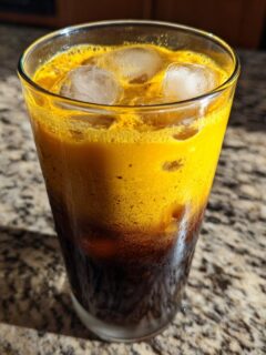 A tall glass containing a layered Honey Turmeric Iced Latte with ice cubes, showing a dark bottom layer and a vibrant yellow top layer.