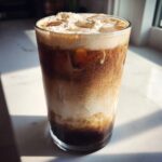 A tall glass of Iced Vanilla Sweet Cream Latte Copycat showing layers of dark coffee, sweet cream mixing, and a thick foam topping.