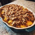 Close-up of an Irresistible Peach Crisp With Fresh Peaches in a baking dish, showing bubbling fruit and a golden crumble topping.