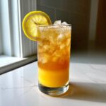 A tall glass filled with a layered Mango Lemonade Kombucha Mocktail, ice, and garnished with a lemon slice.