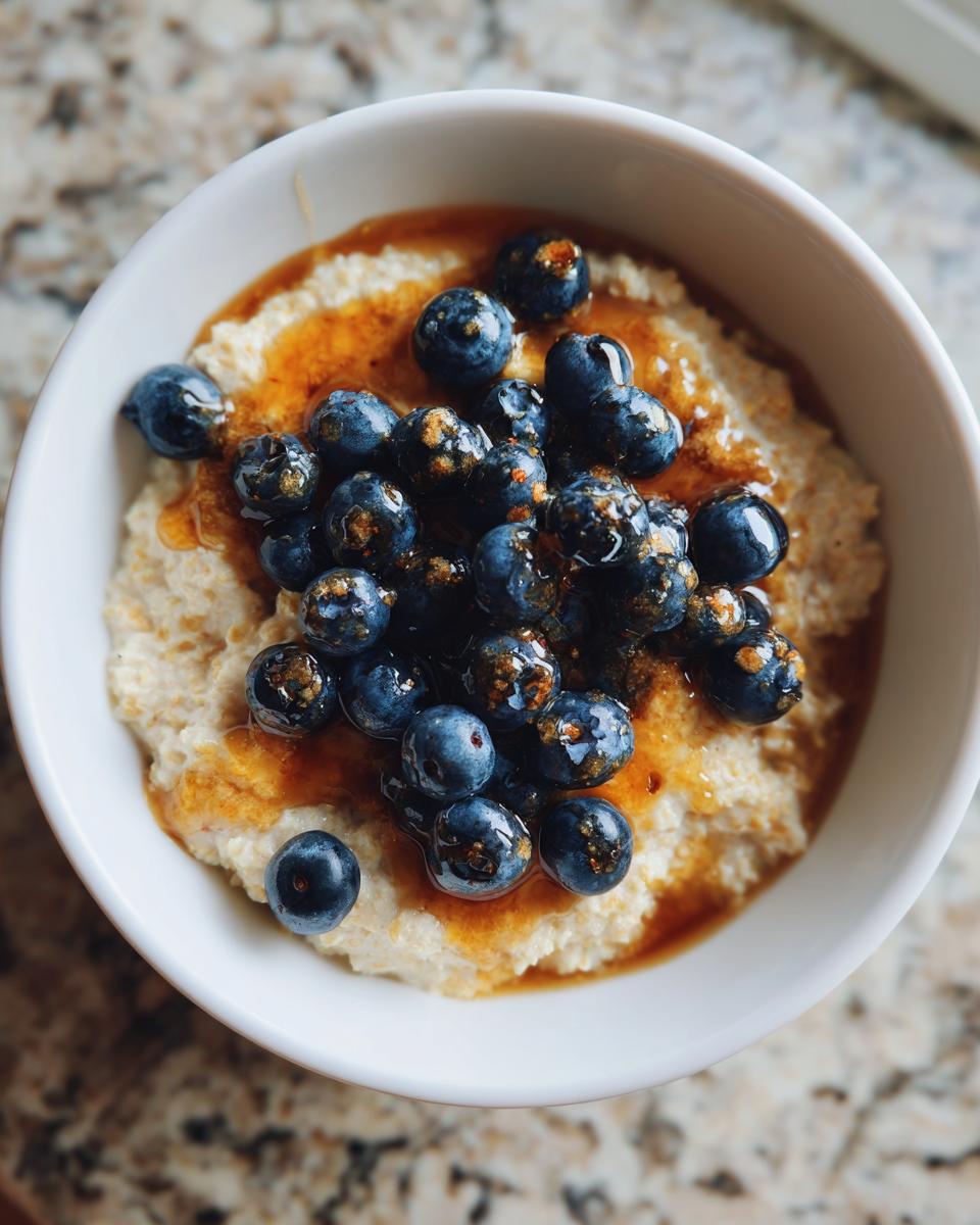 A close-up of a white bowl filled with creamy Maple Blueberry Oatmeal Porridge, topped with fresh blueberries and drizzled with maple syrup.