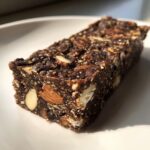 A single, rich, dark No Bake Cacao Date Loaded Energy Bar studded with whole almonds, resting on a white plate.