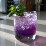 A tall glass filled with ice and vibrant purple Plum Lemonade, topped with fresh mint leaves.