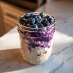 A close-up of Protein Loaded Blueberry Overnight Oats layered in a glass mason jar, topped with fresh blueberries.