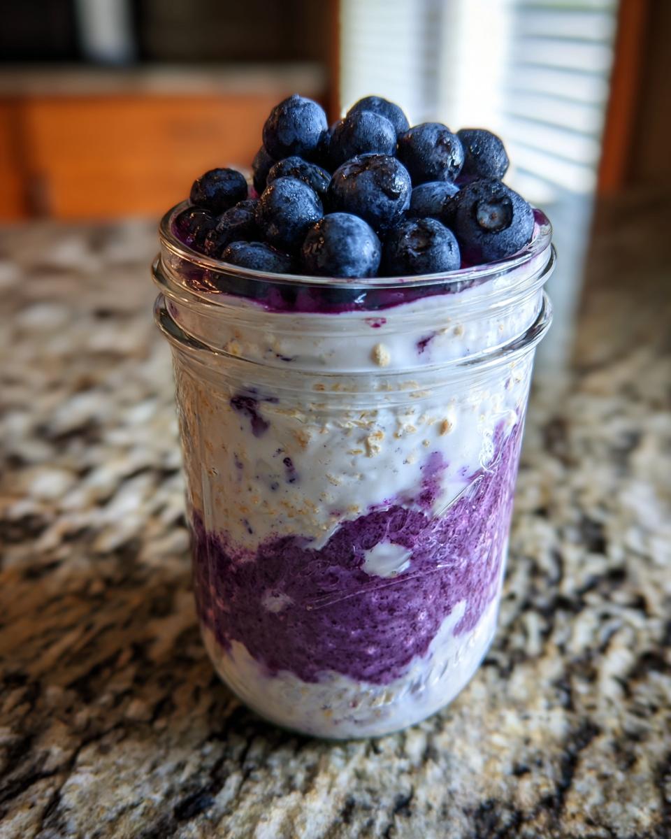 Close-up of Protein Loaded Blueberry Overnight Oats layered with purple blueberry puree in a glass jar, topped with fresh blueberries.