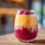 A vibrant two-layered Raspberry Sunrise Smoothie in a glass, topped with fresh raspberries.