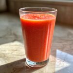 A tall glass filled with a vibrant red Strawberry Mango Papaya Smoothie, catching the sunlight.