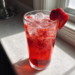 A tall glass filled with a vibrant red Strawberry Pomegranate Mocktail, ice cubes, and garnished with a fresh strawberry slice.