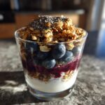 A close-up of a Vegan Breakfast Parfait Blueberry layered in a glass with yogurt, granola, and chia seeds.