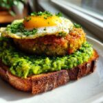 Close-up of Vegan Pesto Egg Avocado Toast featuring bright green pesto, a patty, and a sunny-side-up egg on toasted bread.