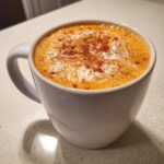 Close-up of a creamy Vegan Pumpkin Chai Latte in a white mug, topped with foam and sprinkled cinnamon.