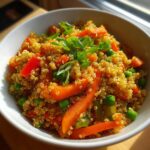 A close-up of a white bowl filled with vibrant Vegan Quinoa Fried Rice, featuring quinoa, carrots, peas, and red bell peppers, topped with green onions.