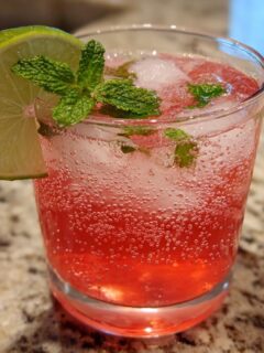 A close-up of a Watermelon Strawberry Lime Mocktail in a glass, garnished with lime and mint, filled with ice and bubbles.