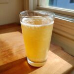 A tall glass filled with fizzy, yellow Apple Cider Vinegar Detox Lemonade sitting on a wooden surface near a window.