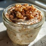 A glass jar filled with Apple Pie Overnight Oats, topped with cream, diced apples, and cinnamon.