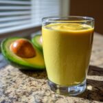 A tall glass of bright yellow Avocado Mango Smoothie next to a halved avocado on a granite countertop.