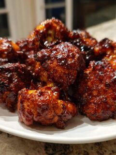 A pile of glistening, saucy BBQ Cauliflower Wings coated in dark, caramelized BBQ sauce, served on a white plate.